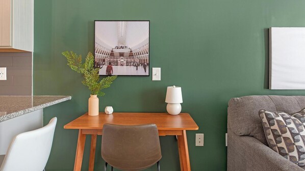 A room with a green wall, a framed photograph, a small table with a chair, a lamp, and a patterned sofa. - Landing | Stylish 1BD, Gym, Yoga Studio (Charlotte)