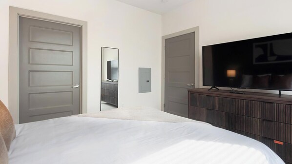 A hotel room with a bed, a TV on a wooden dresser, and a door leading to another room. - Landing | Stylish 1BD, Gym, Yoga Studio (Charlotte)