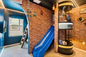 Ein Zimmer mit Backsteinwand, einer blauen Rutsche, einer Leiter und einer Glasvitrine mit diversen Gegenständen.