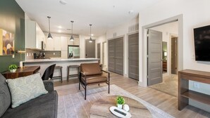 Uma sala de estar moderna com sofá, mesa de centro e uma poltrona. Há uma área de cozinha com geladeira e mesa de jantar. O quarto possui um espelho grande e uma TV montada na parede.