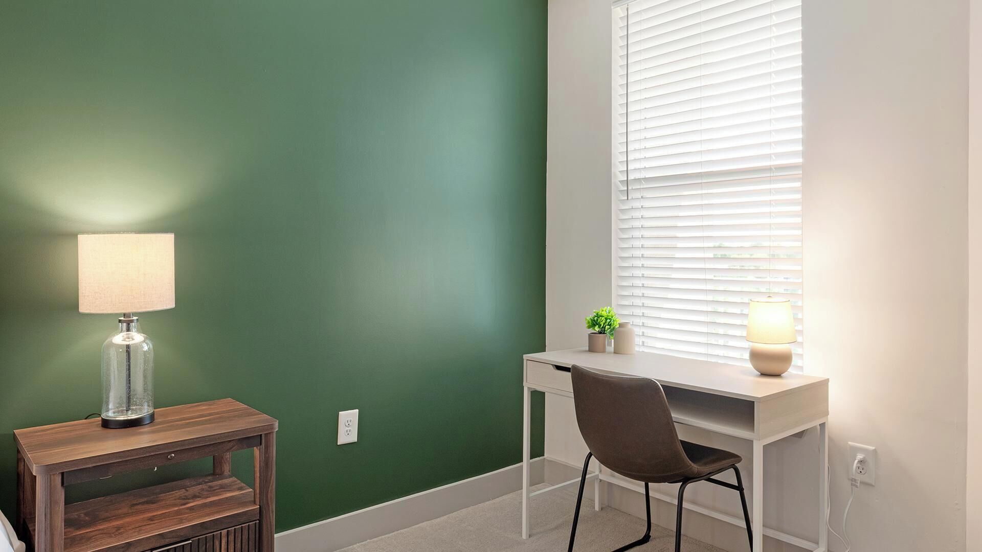 A room with a desk, chair, lamp, and a window with blinds.