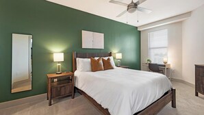 Une chambre à coucher comprenant un lit, deux tables de chevet, un bureau, une chaise, un ventilateur de plafond et une fenêtre avec des stores.