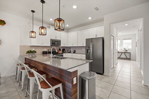 Een moderne keuken met een centraal eiland, barstoelen, roestvrijstalen apparatuur, hanglampen en uitzicht op de aangrenzende woonkamer.