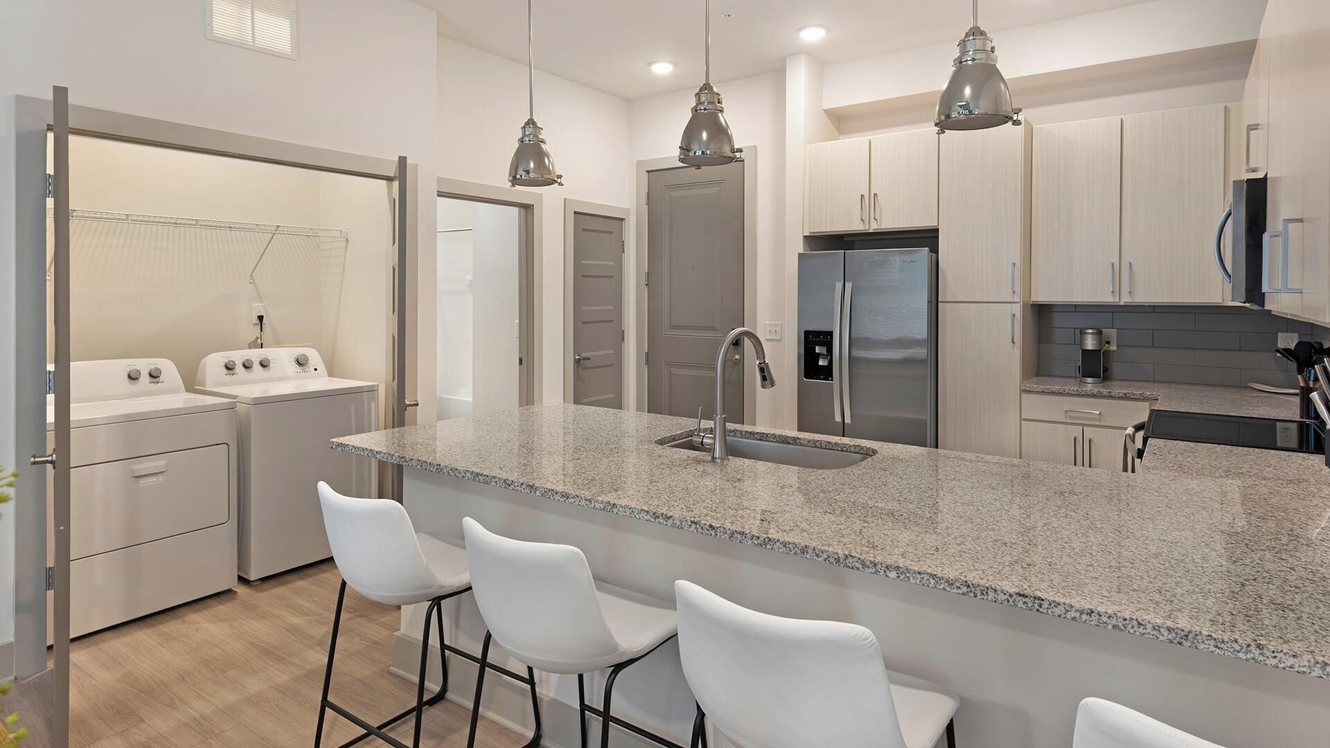 Una cocina moderna con encimeras de granito, electrodomésticos de acero inoxidable y taburetes de bar blancos.