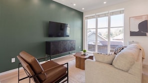 A modern living room with a green wall, a flat-screen TV, a leather chair, a sofa, and a window with a view of houses. - Landing | Stylish 3BD in Dallas (Dallas)