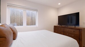 Ein Schlafzimmer mit einem Bett, einem Fernseher auf einer Holzkommode und einem Fenster mit Jalousien.