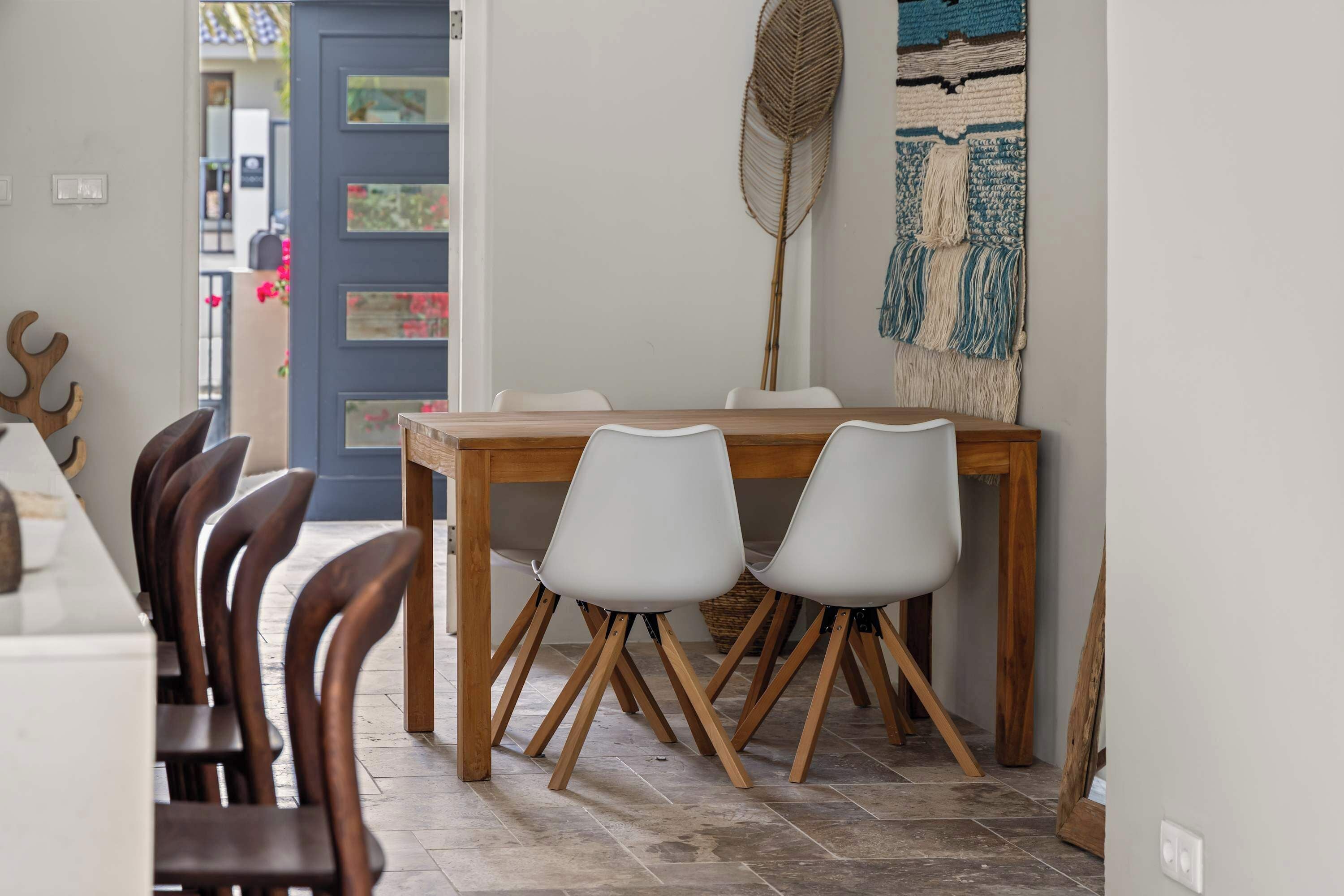 A dining area with a wooden table and white chairs, a blue door, and a wall-mounted fan.