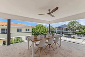 A covered outdoor dining area with a table and chairs, a ceiling fan, and a view of surrounding buildings and greenery.