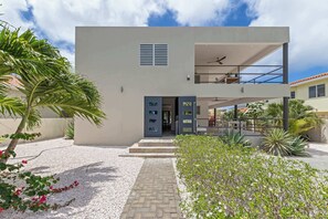 Ein modernes, zweistöckiges Haus mit Flachdach, einem Balkon mit Deckenventilator und einem kleinen Gartenbereich mit Kies und Pflanzen.