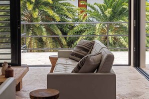 A modern living room with a leather sofa, a round wooden table, and a view of tropical foliage through large windows.