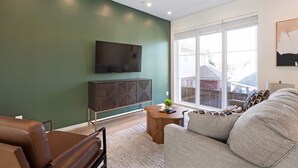 A modern living room with a large flat-screen TV mounted on the wall, a dark wood cabinet below it, a grey sofa, and a small wooden side table with a plant. - Landing | Amazing 3BD in Dallas (Dallas)