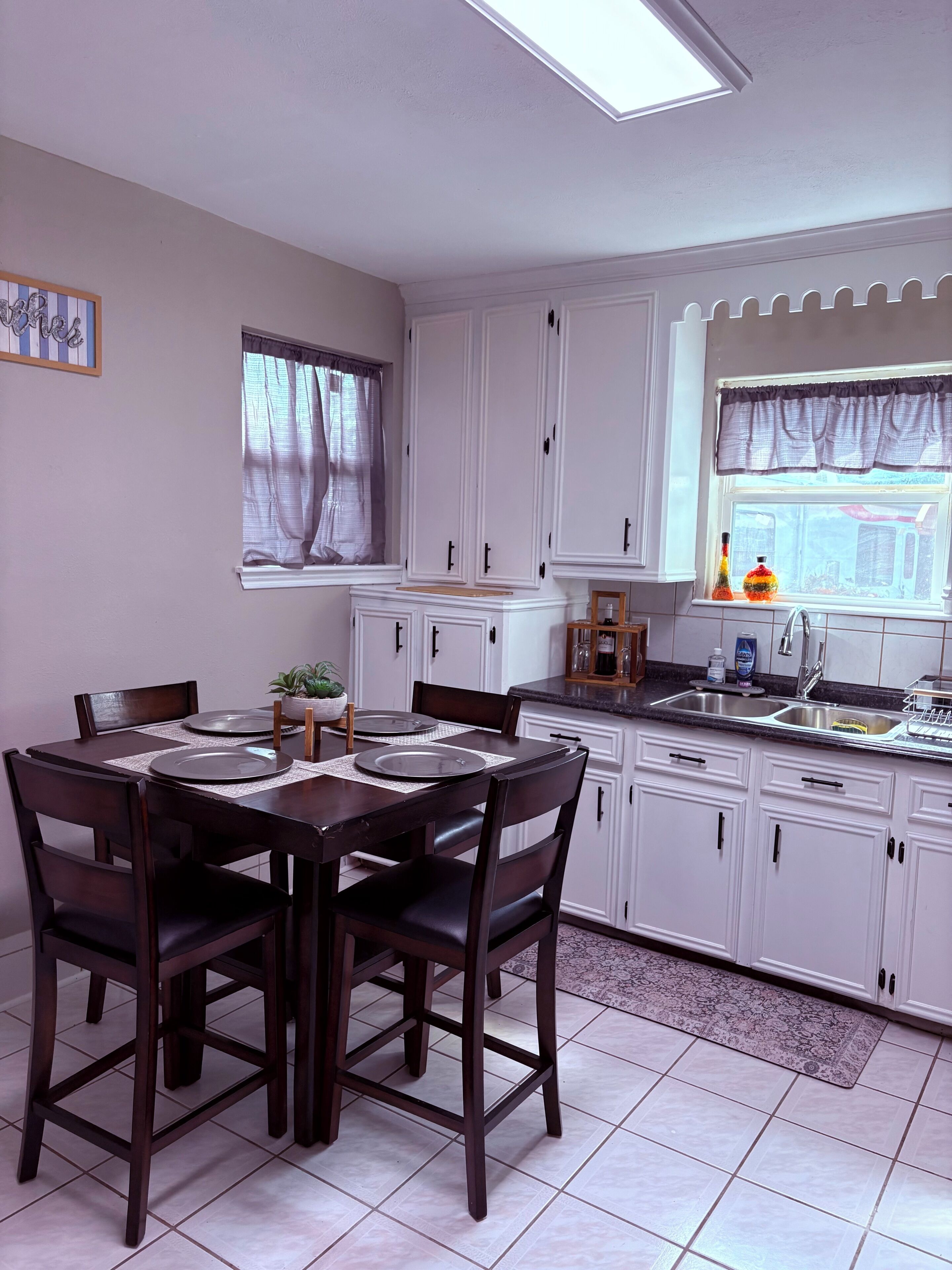 A kitchen with white cabinets, a dining table with chairs, a window with curtains, and a sink.