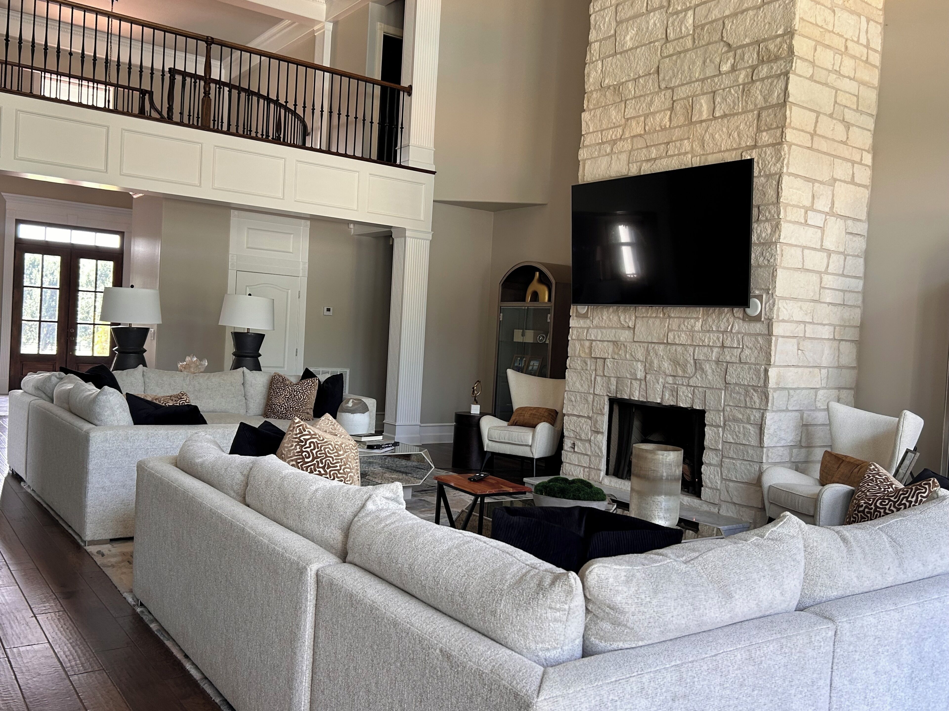 A spacious living room with a sectional sofa, a fireplace, a flat-screen TV mounted on a stone wall, and a balcony with a railing.