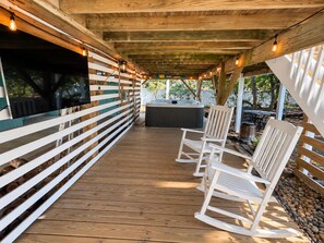 An outdoor deck with wooden flooring, white rocking chairs, a hot tub, and a TV mounted on a wooden wall. - Newly Renovated Family Home in Ocean Sands (Corolla)