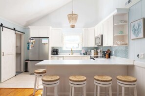 A modern kitchen with white cabinetry, a large island with stools, a hanging light fixture, and a window with a view. - Newly Renovated Family Home in Ocean Sands (Corolla)