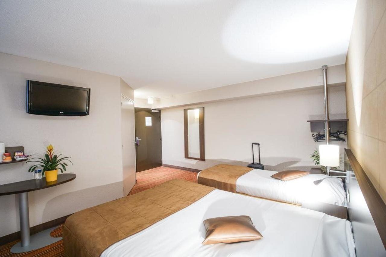 Een hotelkamer met twee bedden, een aan de muur bevestigde televisie, een bureau met een plant en een spiegel.