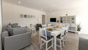 Een moderne woonkamer met een grijze bank, een eettafel met stoelen, een flatscreen televisie en een witte boekenkast met decoratieve objecten.