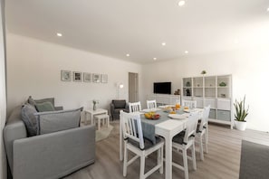 Ein modernes Wohnzimmer mit einem grauen Sofa, einem Esstisch mit Stühlen, einem Flachbildfernseher und einer weißen Regaleinheit mit Dekorationselementen.