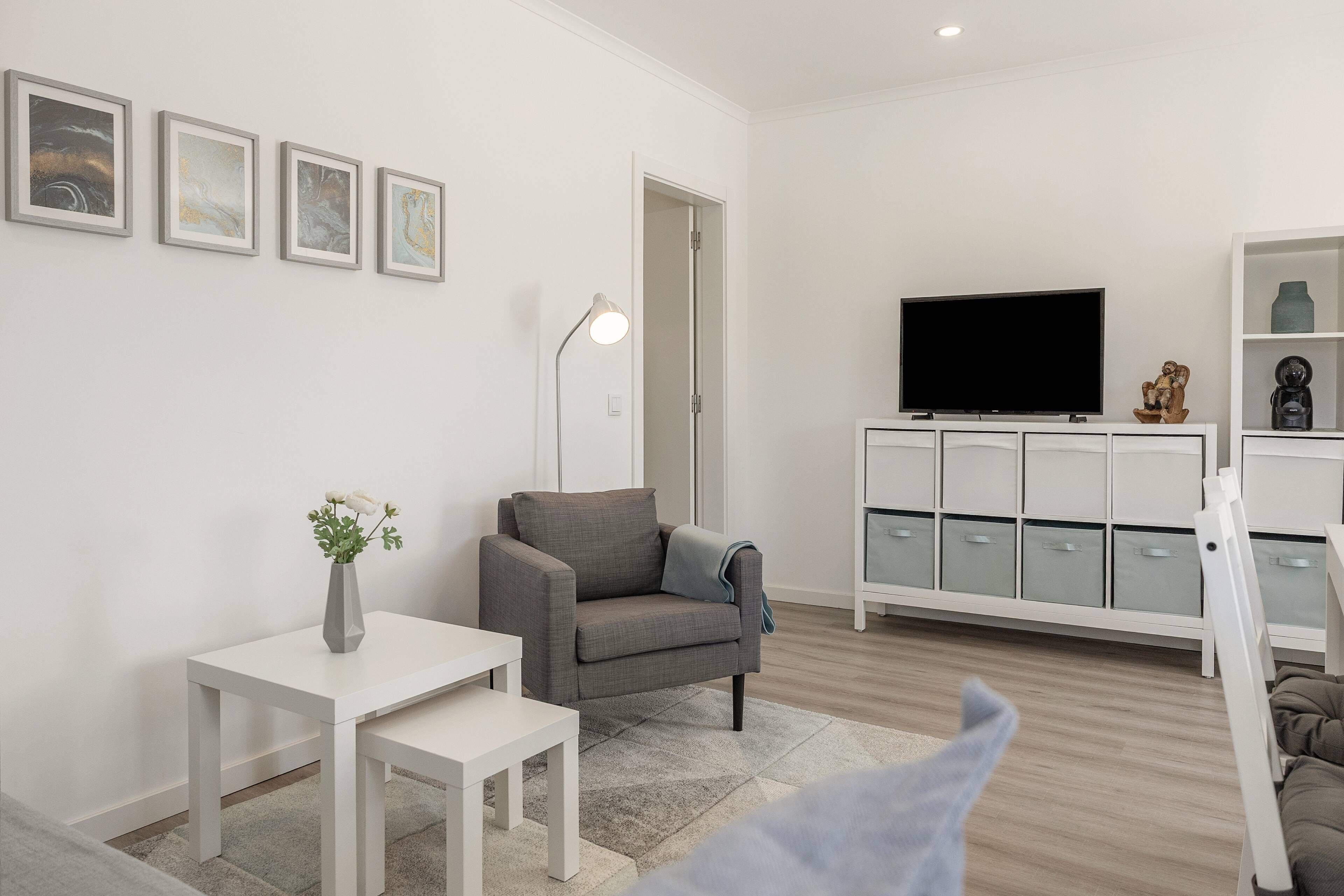 Un salon moderne avec un canapé gris, une table blanche, un téléviseur posé sur un meuble et un buffet doté de tiroirs.