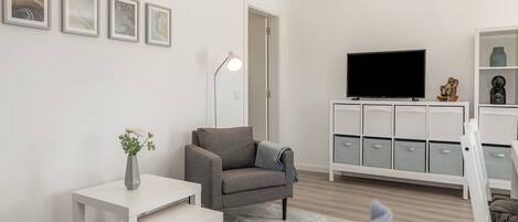 Un salon moderne avec un canapé gris, une table blanche, un téléviseur posé sur un meuble et un buffet doté de tiroirs.