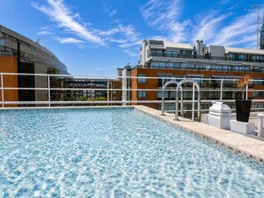 Ein Schwimmbecken mit klarem, blauem Wasser, umgeben von einem weißen Zaun und einem modernen Gebäude mit zahlreichen Fenstern und Balkonen.