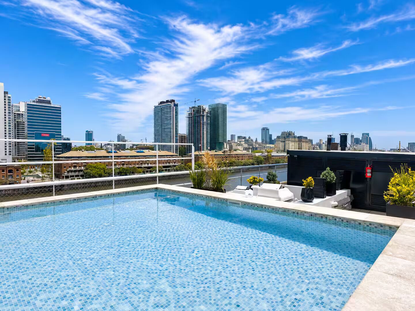 A rooftop pool with a city skyline view, surrounded by a white fence and potted plants.