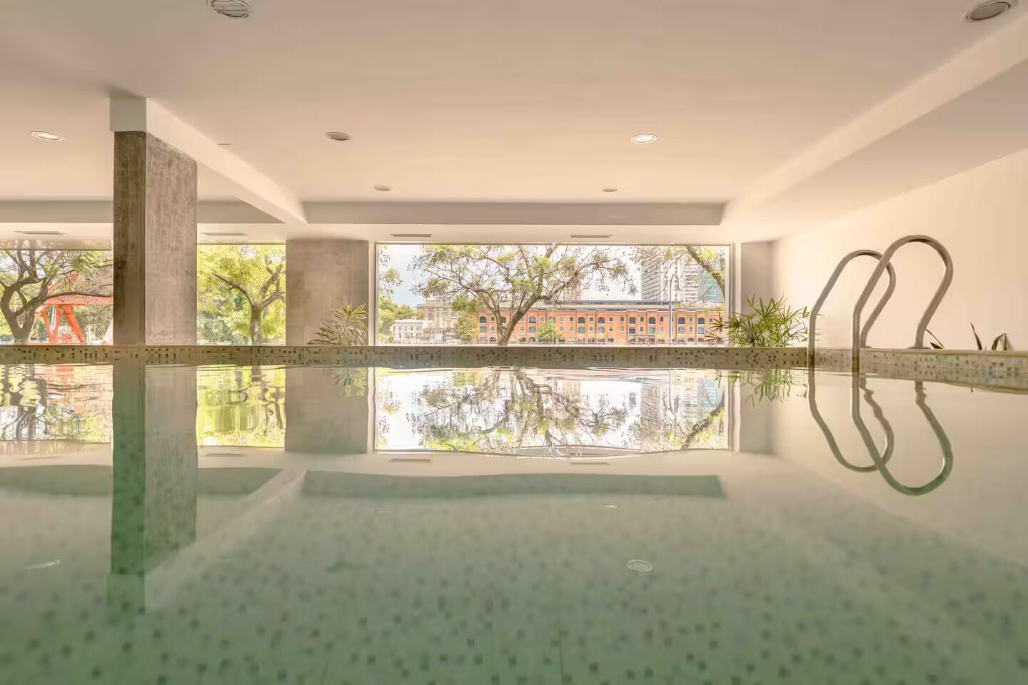 A modern indoor pool with clear reflections, large windows, and a view of a building and trees.