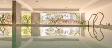 A modern indoor pool with clear reflections, large windows, and a view of a building and trees.