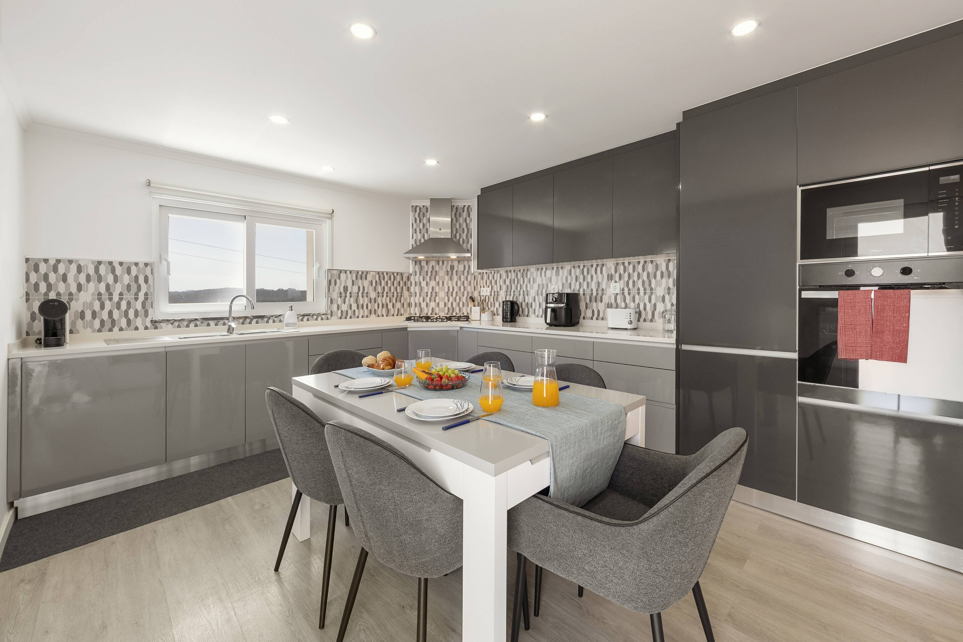A modern kitchen with grey cabinetry, a white countertop, and a dining area with grey chairs.