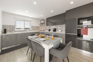 A modern kitchen with grey cabinetry, a white countertop, and a dining area with grey chairs. - Casa Saudade - Vila Serena (Caldas da Rainha)