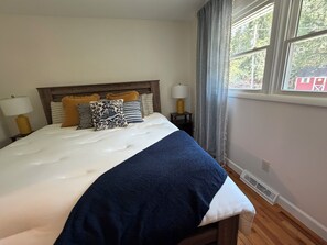 Ein ordentlich bezogenes Bett mit blauer Decke, einem Holz-Kopfteil und mehreren Kissen. Ein Fenster mit Vorhängen und eine Lampe auf einem Nachttisch sind ebenfalls zu sehen.