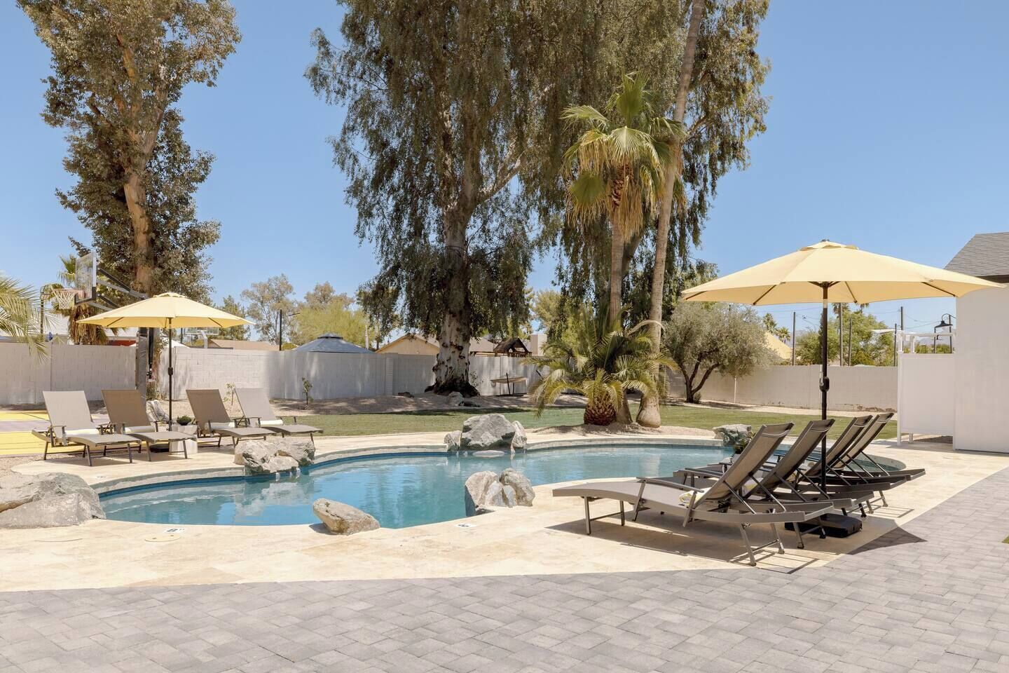 Une piscine entourée d’arbres et d’une clôture blanche, avec des chaises longues et des parasols à proximité.