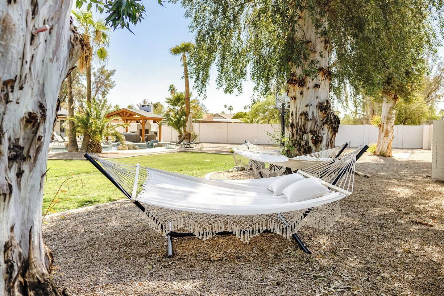 Two hammocks under trees in a backyard with a pool and a gazebo.