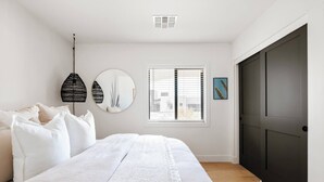 Um quarto com uma cama, um espelho redondo, uma janela com persianas e uma porta.