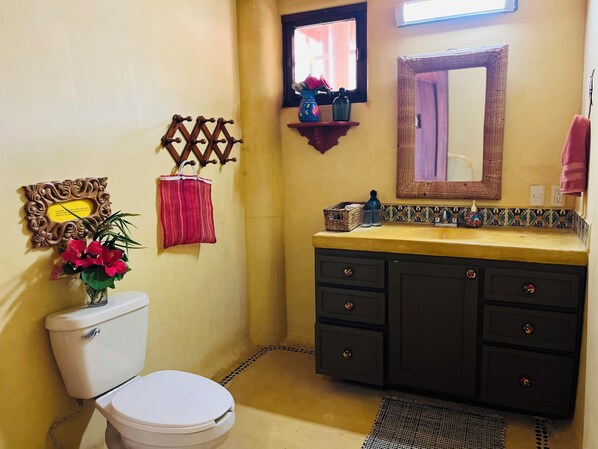 A bathroom with a toilet, a sink with a mirror, a decorative wall hanging, and a small shelf with items.