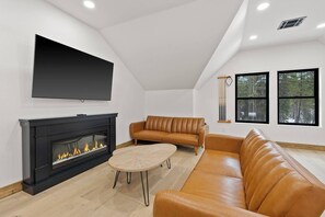 A modern living room with a fireplace, leather sofas, a TV mounted on the wall, and large windows. - Huckleberry Peak Villa -Glacies Montana-Game Room (Hungry Horse)