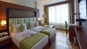 A hotel room with two beds, a desk, a chair, a television, and a window with curtains. - Shri Ram Empire (Jodhpur)