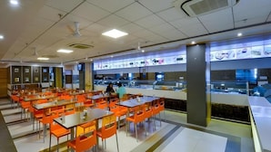 A cafeteria with multiple tables and chairs, a counter with food displays, and ceiling fans. - Shri Ram Empire (Jodhpur)
