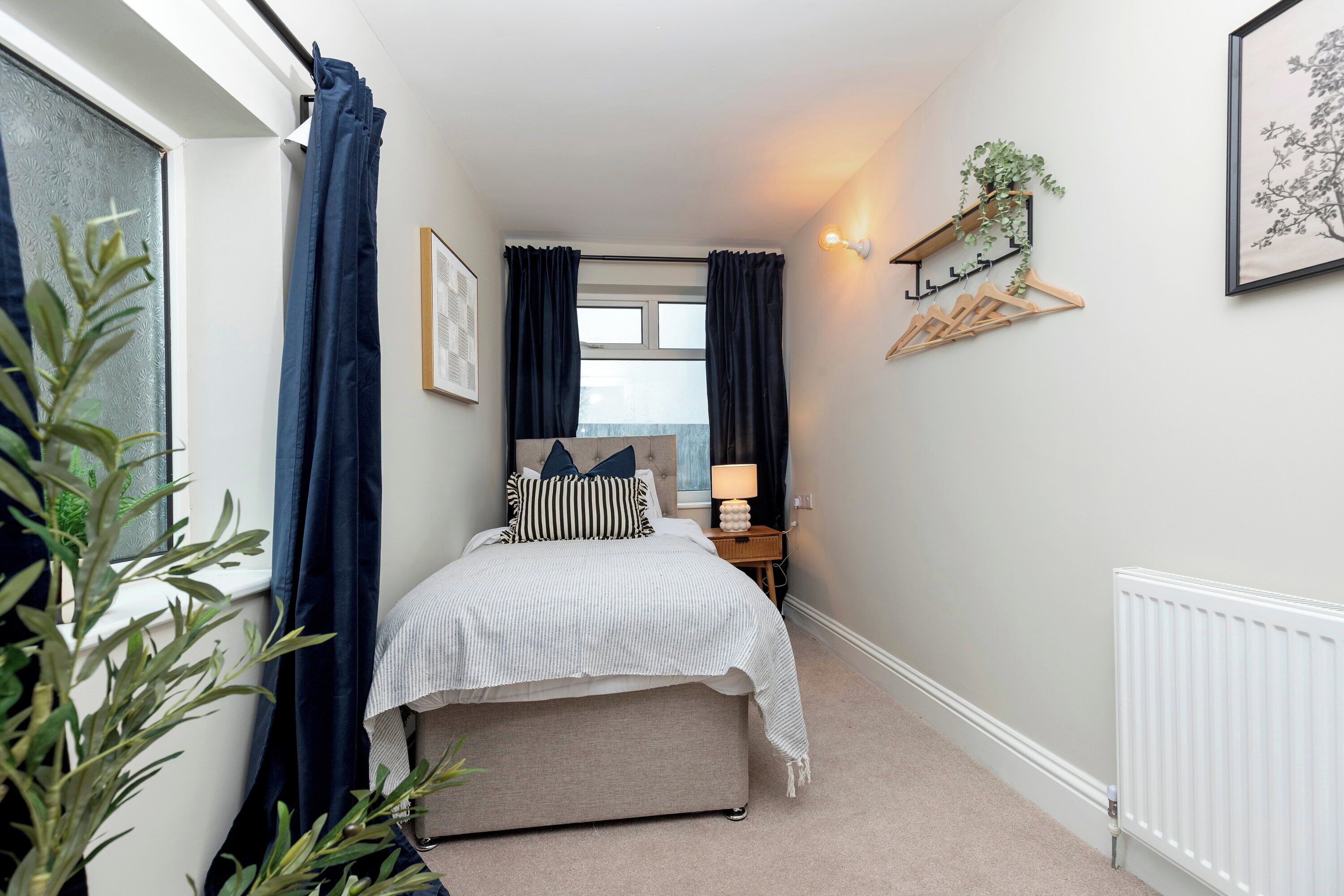 Un dormitorio con cama, mesita de noche con lámpara, ventana con cortinas, radiador y estante en la pared con una planta.
