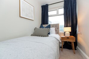 A neatly made bed with a striped pillow, a bedside table with a lamp, and a window with curtains. - Gateshead 3BR Stay | Modern & Contractor Friendly (Ryton)