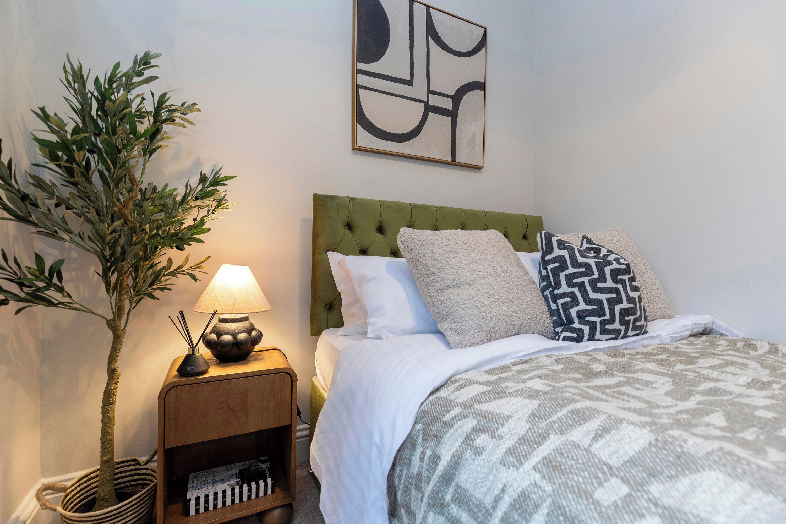 A neatly arranged bedroom with a green upholstered headboard, a patterned bedspread, and a wall-mounted abstract painting.