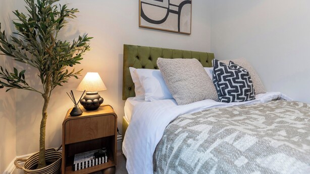 Une chambre à coucher bien rangée, avec une tête de lit verte en tissu, une couvre-lit à motifs et un tableau abstrait accroché au mur.