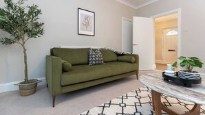 Un salon avec un canapé vert, une plante en pot, une estampe botanique encadrée et un tapis à motifs.
