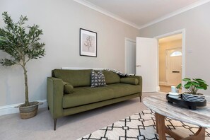 Ein Wohnzimmer mit einem grünen Sofa, einem Blumentopf, einem gerahmten Pflanzenprint und einem gemusterten Teppich.