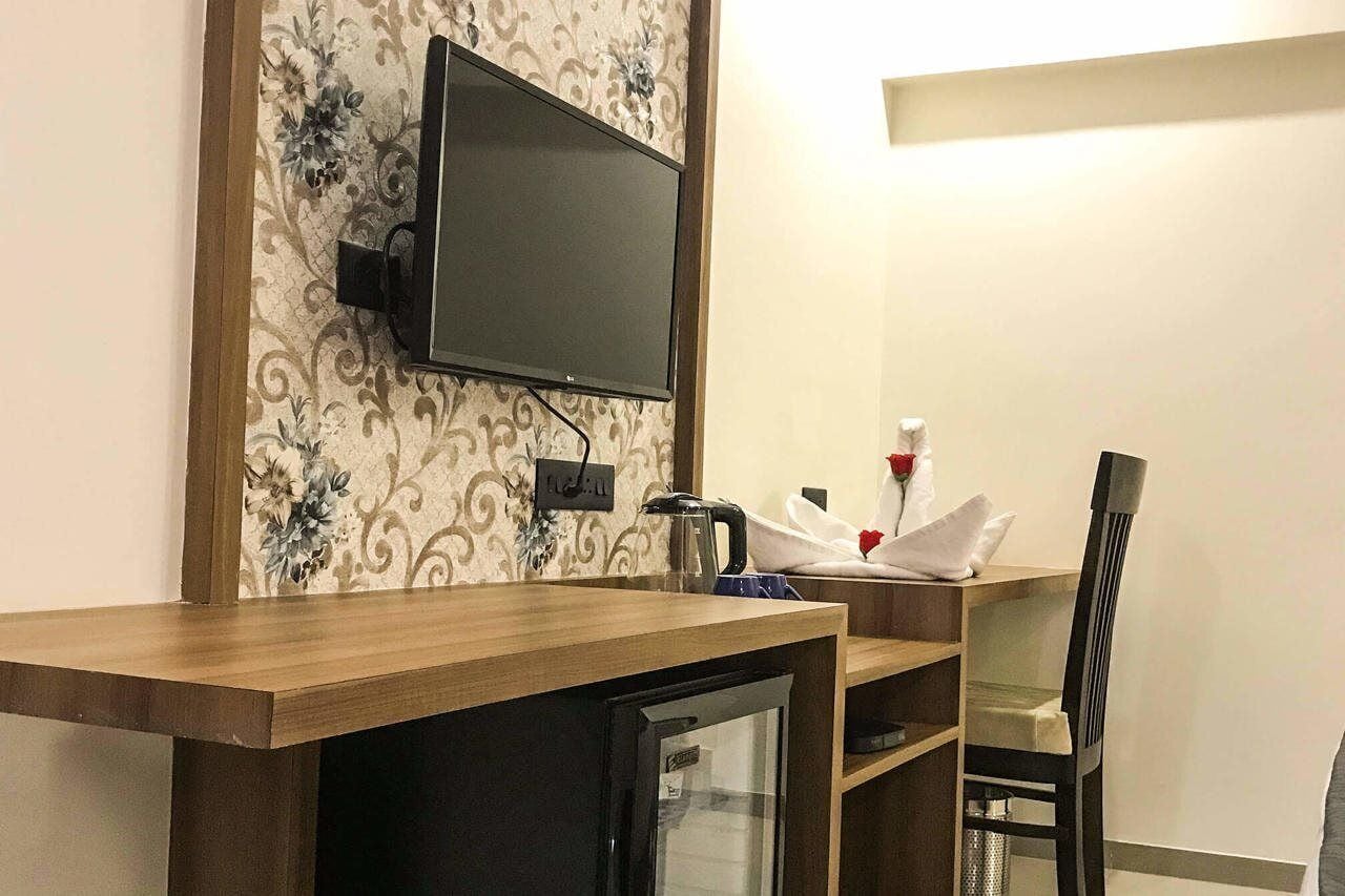 A hotel room with a wooden desk, a television mounted on the wall, a small refrigerator, and a dining area with a chair and table.