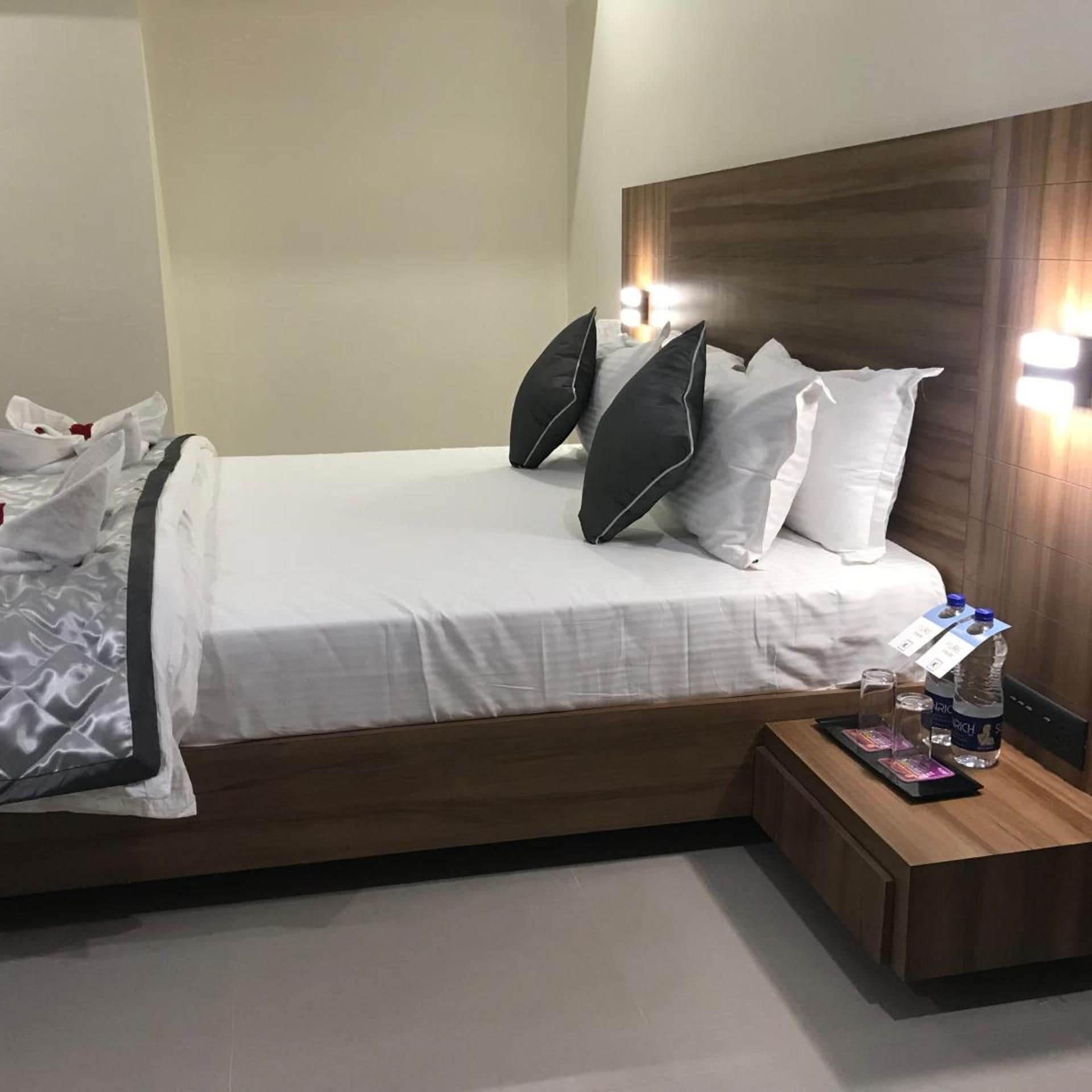A hotel room with a bed, pillows, a bedside table with a water bottle, and a tray with a card and glasses.