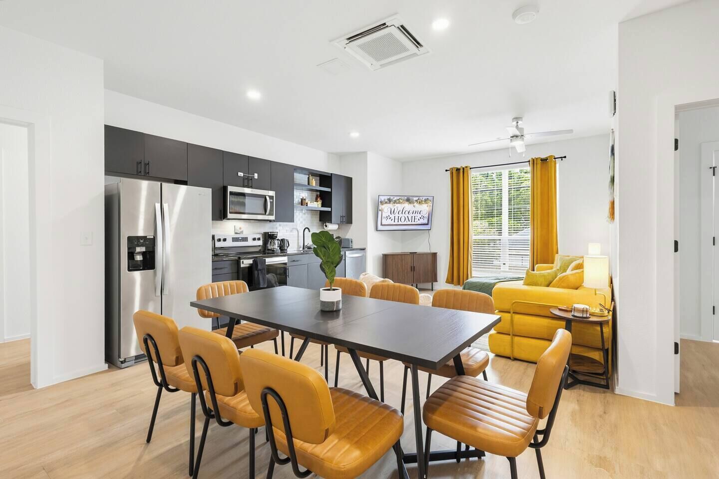A modern kitchen with stainless steel appliances, a dining area with wooden chairs, and a yellow sofa in a bright living room.