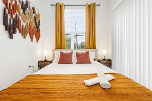 A hotel room with a bed, two bedside tables, a window with curtains, and a wall with hanging items. - Walk to Bishop Arts | Coffee & Dining Nearby Walk! (Dallas)