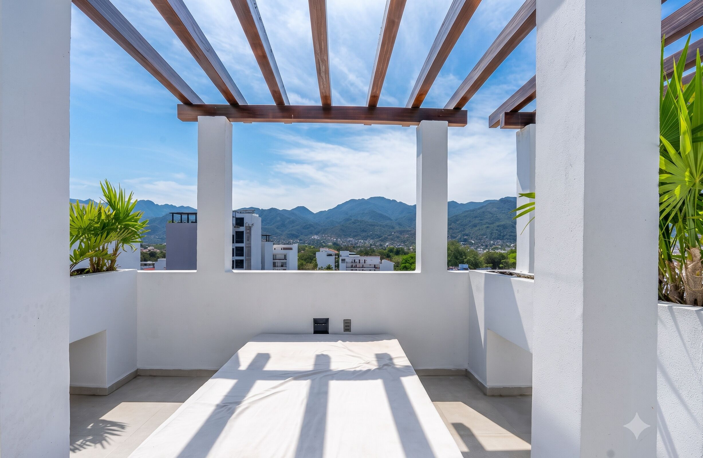 Un balcón con vistas a montañas y edificios, una pérgola de madera y plantas en macetas.