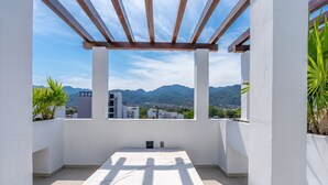 Un balcón con vistas a montañas y edificios, una pérgola de madera y plantas en macetas.
